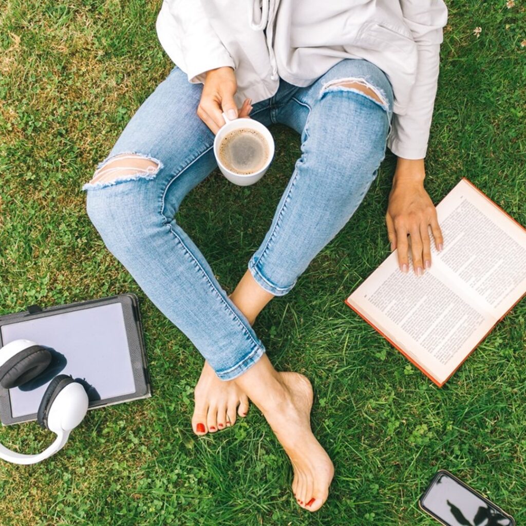 Pessoa estudando nas férias ao ar livre com um livro, tablet, fones de ouvido e uma xícara de café, sentada na grama.