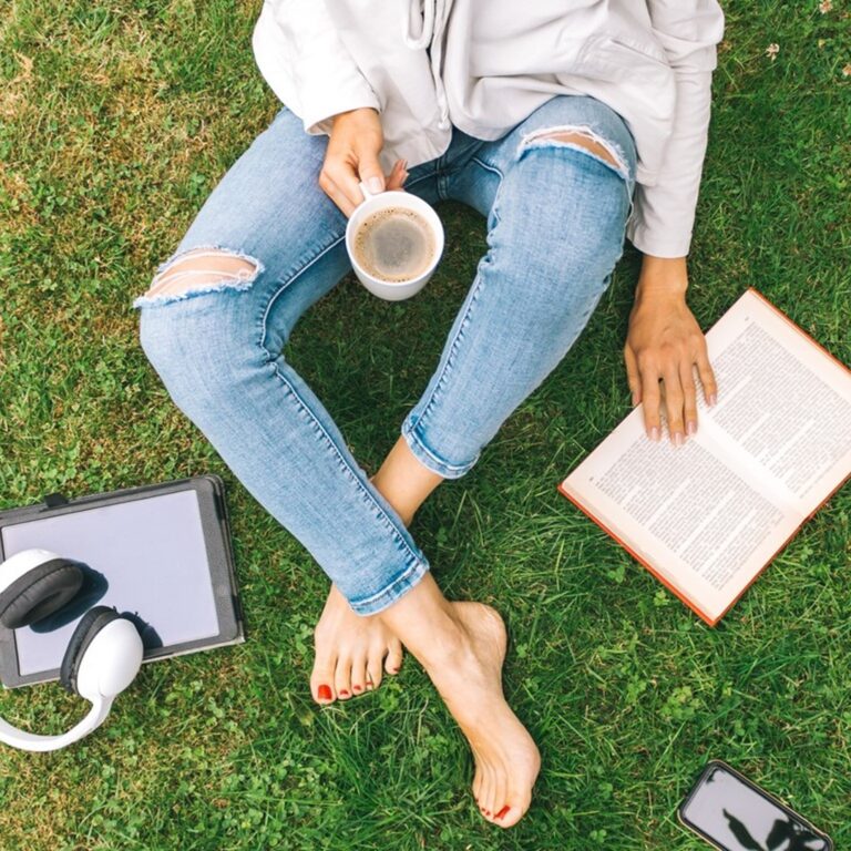 Pessoa estudando nas férias ao ar livre com um livro, tablet, fones de ouvido e uma xícara de café, sentada na grama.
