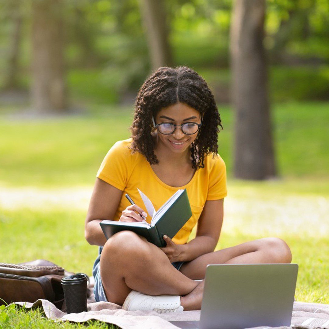 Jovem estudando ao ar livre durante as férias escolares, sentada na grama com um notebook e um caderno.