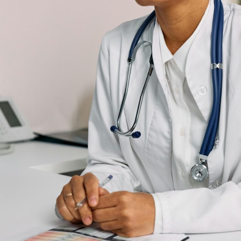 Imagem de um profissional de medicina vestindo um jaleco branco e um estetoscópio em referência aos estudantes que desejam passar em medicina. Ao lado, vemos uma mesa com teclado, mouse, telefone e pastas com papéis.
