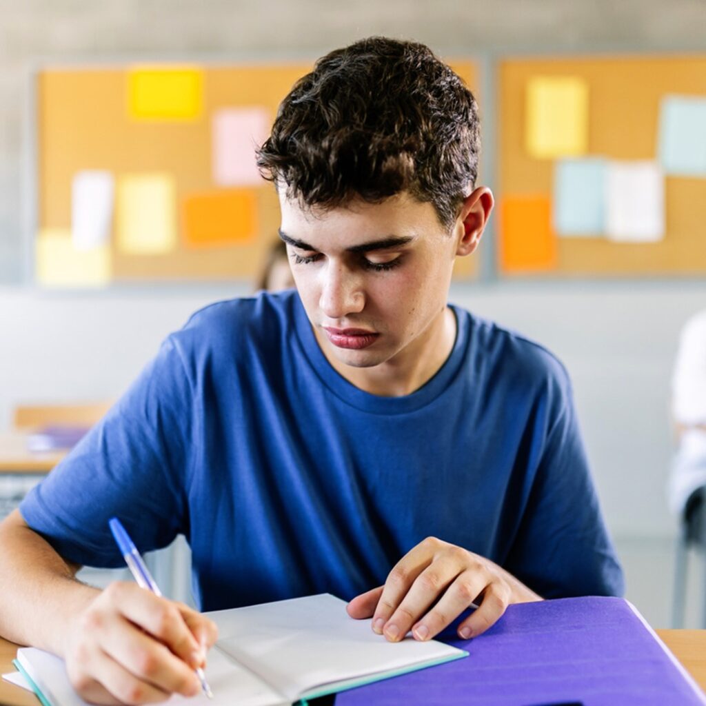 Estudante focado escrevendo em seu caderno, treinando temas de redação para o Enem. A imagem destaca a importância de praticar a escrita e estar preparado para os possíveis tópicos abordados no exame.