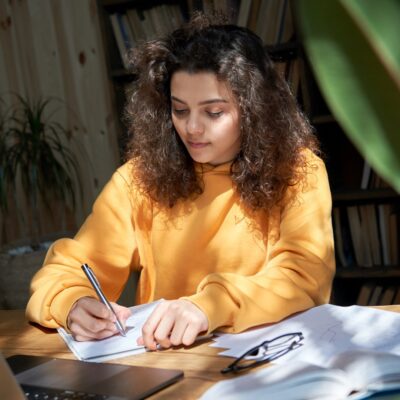 Estudante criando uma rotina de férias em casa, escrevendo e estudando em um caderno ao lado de um laptop.