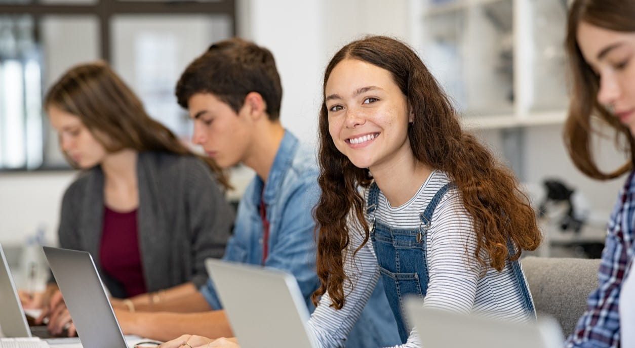 Grupo de estudantes em uma sala de aula usando notebooks, com uma aluna sorrindo para a câmera, ilustrando o conceito de estudo de gramática, como o tema 'o que é adjetivo'.