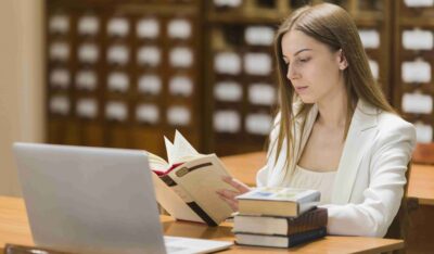 Mulher jovem sentada em uma biblioteca, lendo um livro enquanto estuda Direito em frente a um laptop, com uma pilha de livros ao lado.