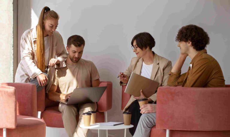 Grupo de quatro pessoas, três homens e uma mulher, sentados em poltronas cor-de-rosa, discutem em um ambiente iluminado. Um deles usa um laptop, enquanto os outros observam e participam da conversa. Todos estão cursando a faculdade de Psicologia.