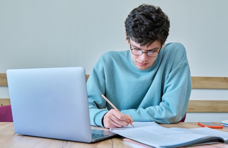 Estudante concentrado estudando e fazendo anotações no caderno com notebook ao lado sobre o Tema da Redação do Enem.