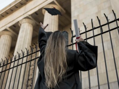 Mulher vestida com beca preta vista de costas, jogando seu chapéu de formatura ao ar em frente a um edifício de colunas, segurando um diploma enrolado com fita vermelha na outra mão.