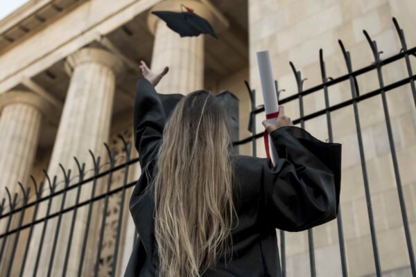 Mulher vestida com beca preta vista de costas, jogando seu chapéu de formatura ao ar em frente a um edifício de colunas, segurando um diploma enrolado com fita vermelha na outra mão.
