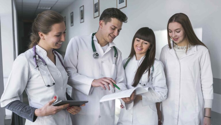 Grupo de jovens estudantes de Medicina, usando jalecos brancos e estetoscópios, analisando um documento em ambiente hospitalar.