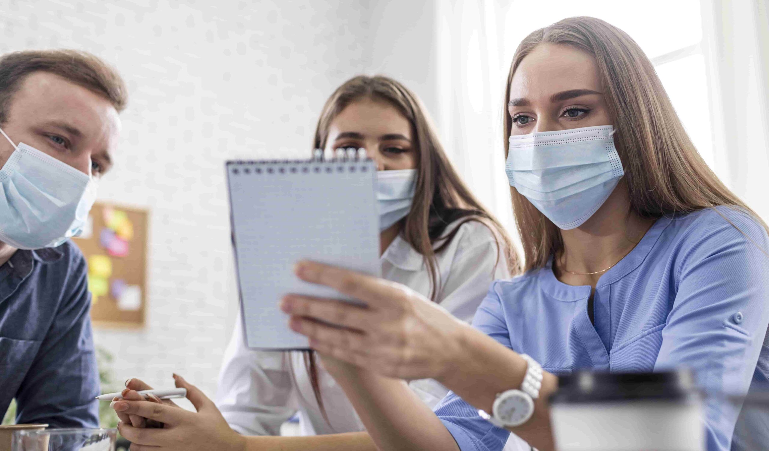 Três estudantes do curso de Enfermagem usando máscaras de proteção participam de uma reunião colaborativa em um ambiente de escritório, analisando anotações em um caderno.