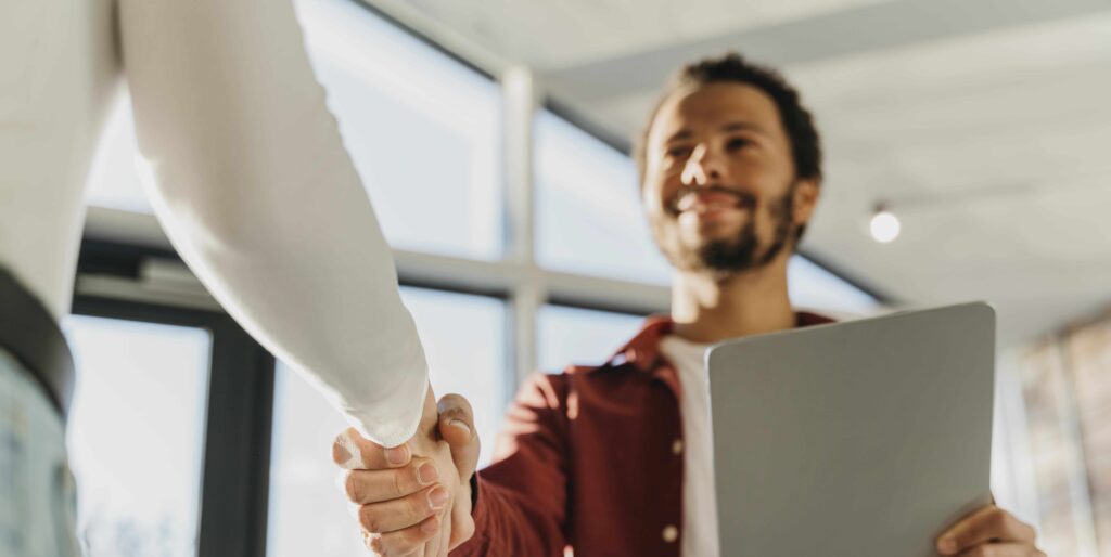 Um homem sorridente segurando um tablet enquanto aperta a mão de outra pessoa, simbolizando uma entrevista de emprego ou fechamento de um acordo profissional em um ambiente corporativo.