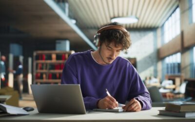 Estudante concentrado em biblioteca, revisando a lista de obras literárias obrigatórias da Fuvest com fones de ouvido e materiais de estudo.