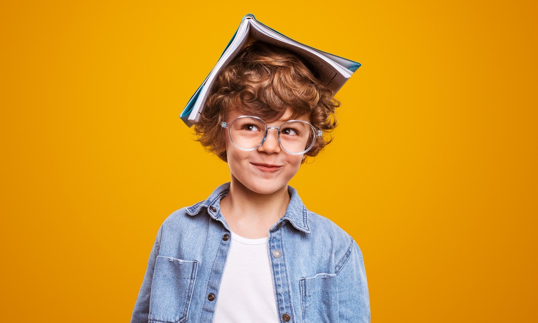 Jovem estudante com expressão divertida e um livro na cabeça, simbolizando aprendizado sobre pronomes em um fundo amarelo vibrante.