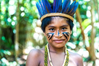 Criança indígena sorrindo na floresta, usando cocar de penas azuis, colares de miçangas e pintura facial tradicional. Identificar traços da cultura indígena ajuda a valorizar suas tradições e história.