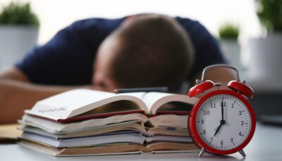 Relógio vermelho marcando horário ao lado de livros e estudante descansando, ilustrando a busca pelo horário ideal para estudar e maximizar a produtividade.