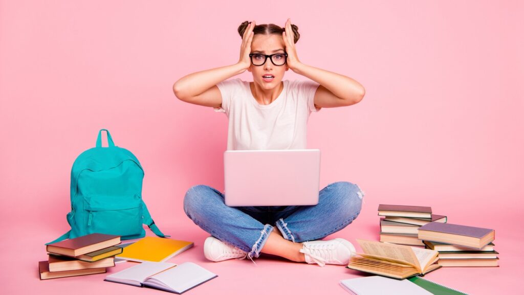 Estudante estressada com notebook e livros ao redor, representando a pressão e desafios de estudar para o vestibular e se preparar para a prova.