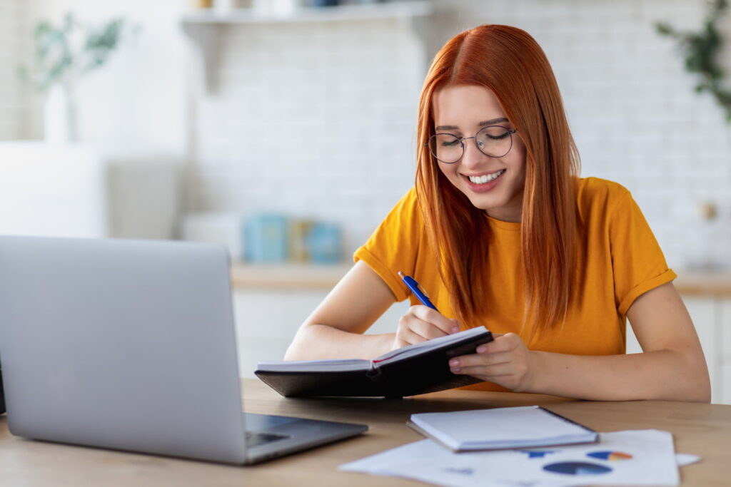 Estudante sorridente escrevendo em caderno enquanto estuda com notebook em casa, representando o tema “vale a pena estudar” para alcançar bons resultados.