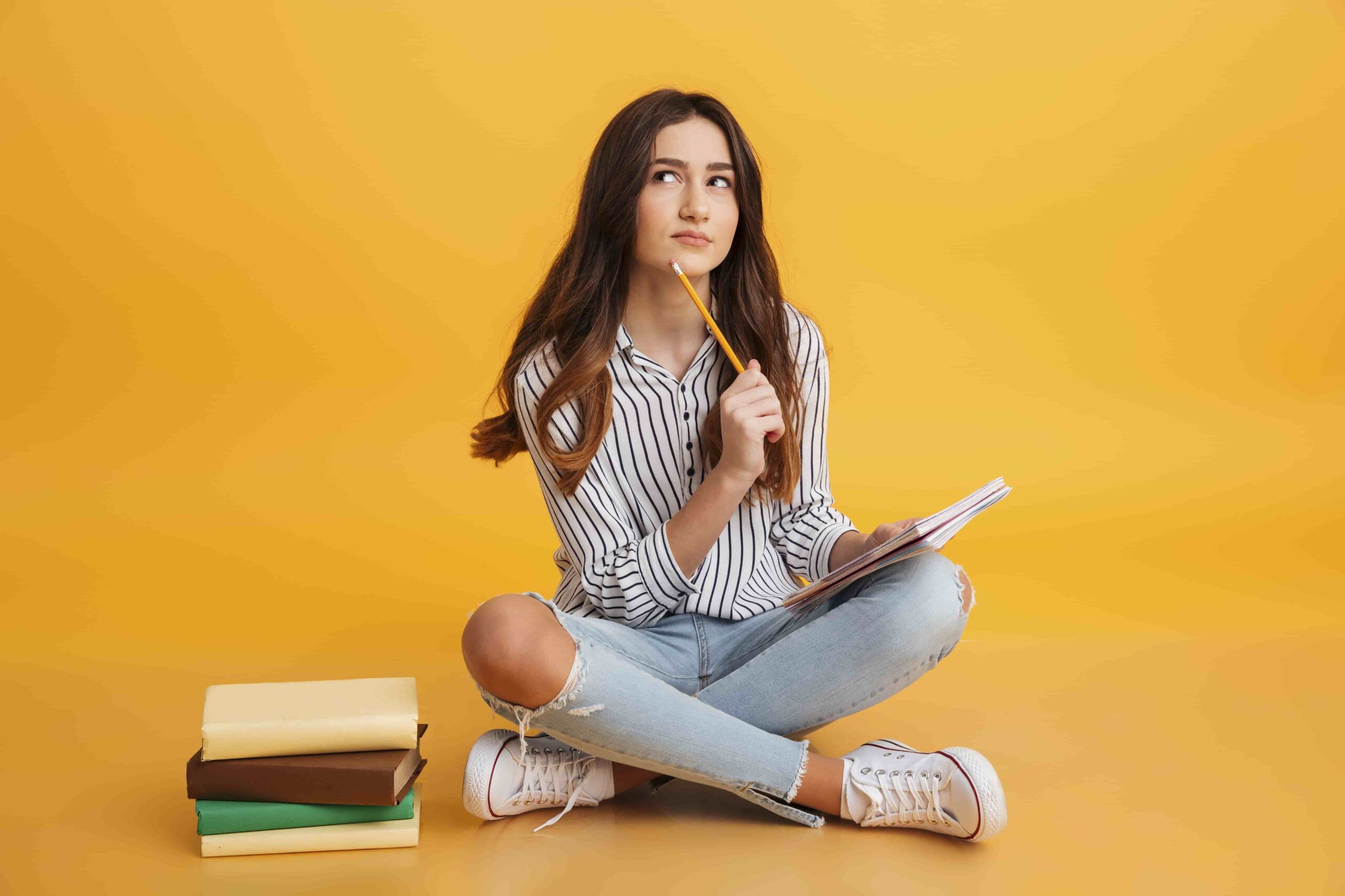 Jovem pensando enquanto segura caderno e lápis, ao lado de uma pilha de livros, simbolizando dúvidas sobre "Como usar o resultado do Enem além do Sisu" para escolher a melhor forma de ingresso.