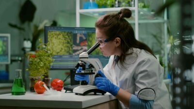 Estudante de Ciências Biológicas analisando amostras vegetais em laboratório, utilizando microscópio e equipamentos de pesquisa, com tomates e plantas ao fundo.