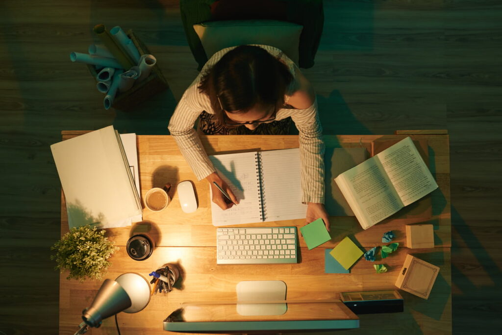 Estudante concentrada escreve em caderno ao lado de livros, computador e post-its, em ambiente iluminado e organizado para estudar em casa com foco e produtividade.