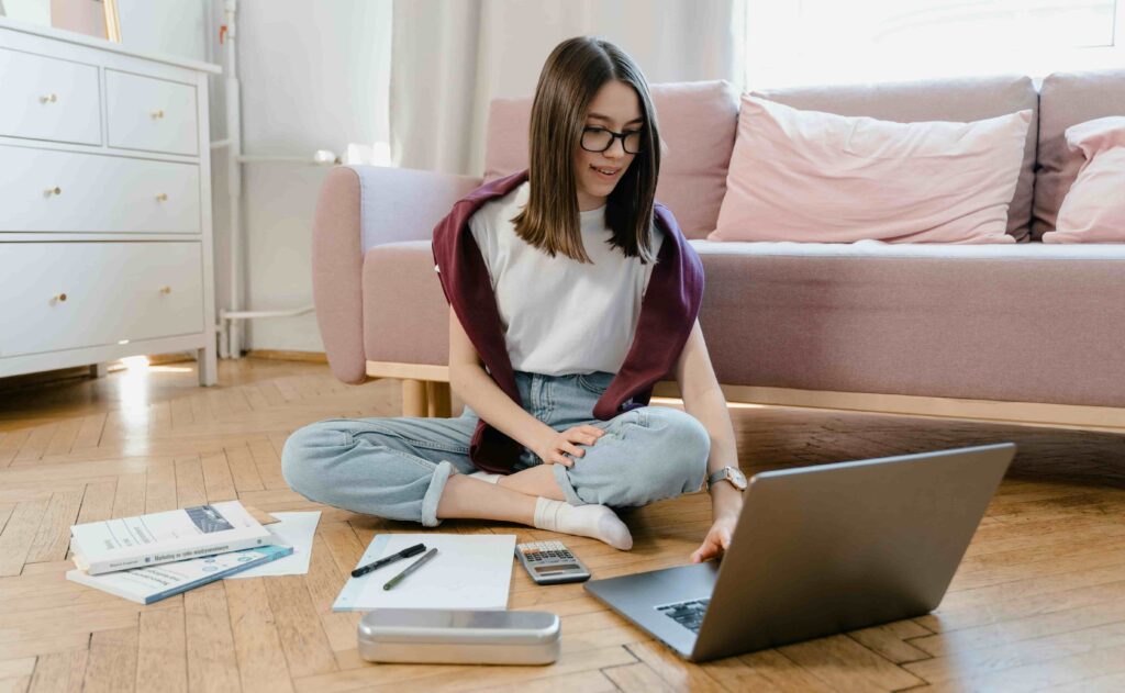 Estudante sentada no chão com um notebook, livros e materiais de estudo, representando o uso de sites para estudar de graça e se preparar para provas do Enem e vestibulares.