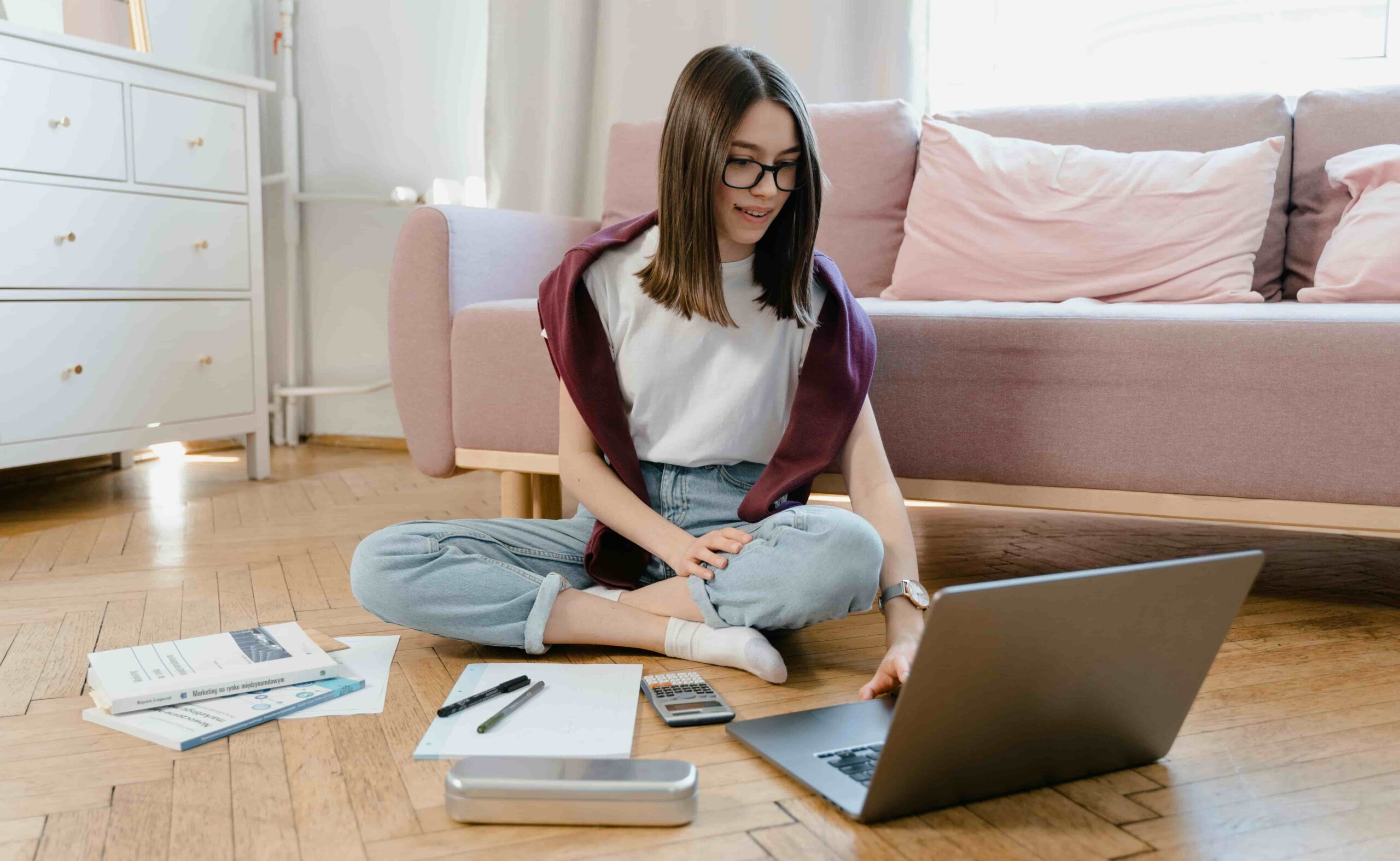 Estudante sentada no chão com um notebook, livros e materiais de estudo, representando o uso de sites para estudar de graça e se preparar para provas do Enem e vestibulares.