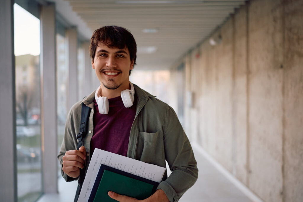 Estudante sorridente em corredor universitário com materiais de estudo, representando o período de inscrições Sisu 2026 e o acesso a vagas no ensino superior.