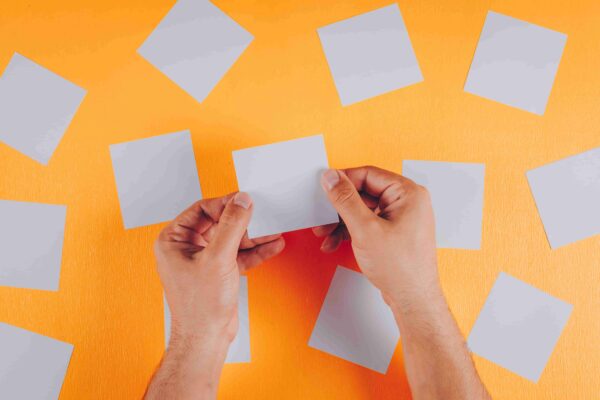 Mãos segurando cartões em branco sobre uma superfície laranja, ilustrando o que são flashcards e como essa técnica de estudo pode ajudar na memorização, revisão e aprendizagem de conteúdos para provas e vestibulares.