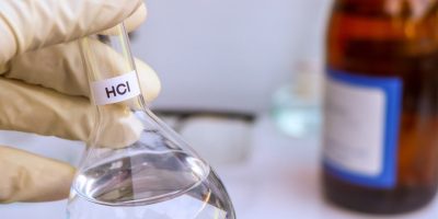 Frasco de laboratório contendo ácido muriático sendo segurado por uma mão com luva, com garrafas e equipamentos de laboratório ao fundo.