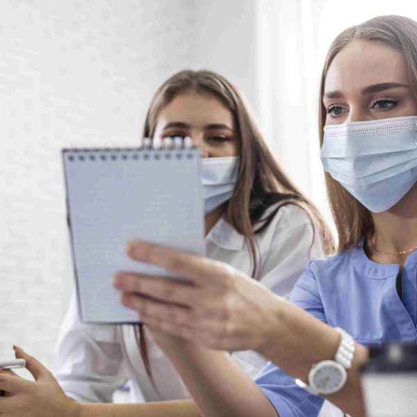 Três estudantes do curso de Enfermagem usando máscaras de proteção participam de uma reunião colaborativa em um ambiente de escritório, analisando anotações em um caderno.