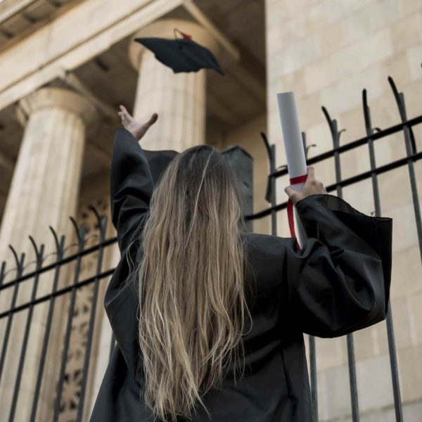 Mulher vestida com beca preta vista de costas, jogando seu chapéu de formatura ao ar em frente a um edifício de colunas, segurando um diploma enrolado com fita vermelha na outra mão.
