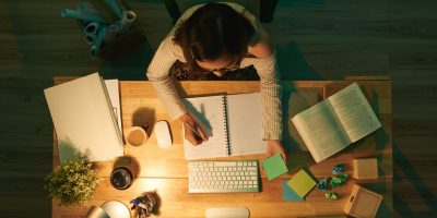 Estudante concentrada escreve em caderno ao lado de livros, computador e post-its, em ambiente iluminado e organizado para estudar em casa com foco e produtividade.