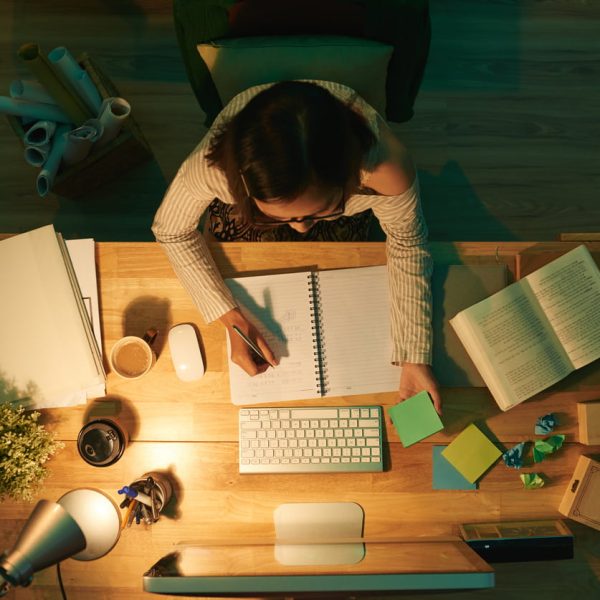 Estudante concentrada escreve em caderno ao lado de livros, computador e post-its, em ambiente iluminado e organizado para estudar em casa com foco e produtividade.