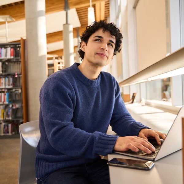 Estudante digitando no laptop em biblioteca iluminada, representando o momento de dúvida sobre a grafia correta de palavras como “encher ou enxer”.
