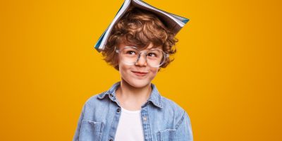 Jovem estudante com expressão divertida e um livro na cabeça, simbolizando aprendizado sobre pronomes em um fundo amarelo vibrante.