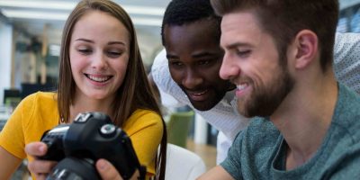 Três estudantes de Publicidade e Propaganda analisam juntos o visor de uma câmera. A imagem transmite colaboração, criatividade e entusiasmo, com foco em projetos de fotografia ou marketing visual.