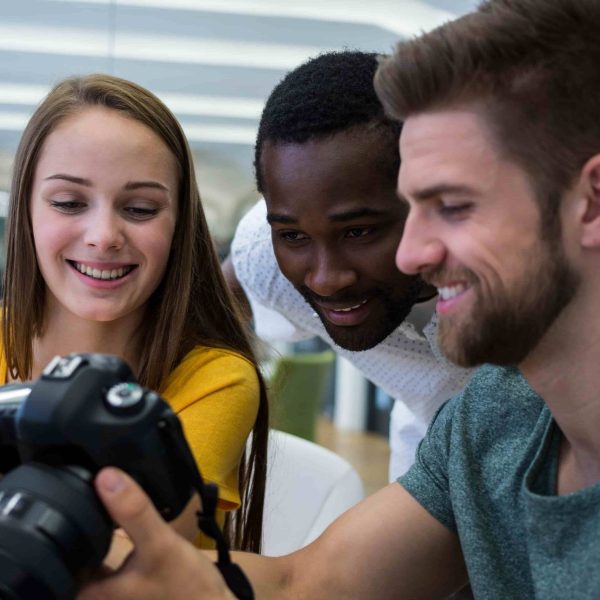 Três estudantes de Publicidade e Propaganda analisam juntos o visor de uma câmera. A imagem transmite colaboração, criatividade e entusiasmo, com foco em projetos de fotografia ou marketing visual.