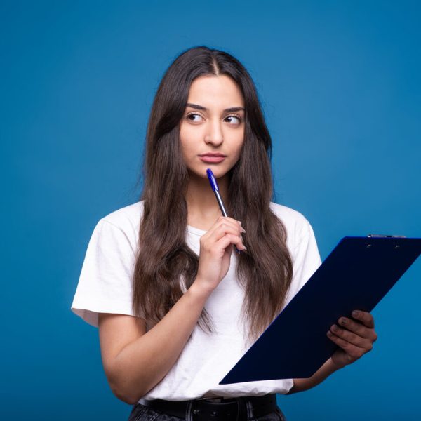 Jovem segurando prancheta e caneta enquanto pensa, ilustrando a dúvida gramatical sobre “sede ou cede”.