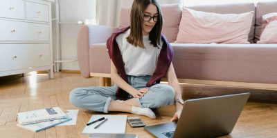 Estudante sentada no chão com um notebook, livros e materiais de estudo, representando o uso de sites para estudar de graça e se preparar para provas do Enem e vestibulares.