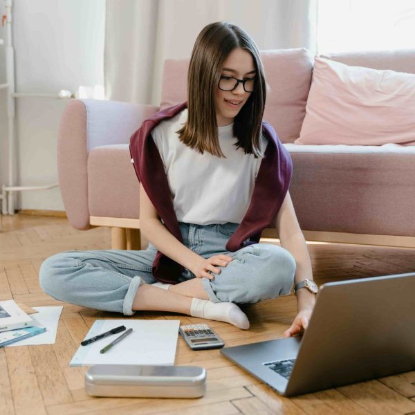 Estudante sentada no chão com um notebook, livros e materiais de estudo, representando o uso de sites para estudar de graça e se preparar para provas do Enem e vestibulares.