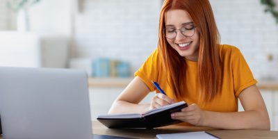 Estudante sorridente escrevendo em caderno enquanto estuda com notebook em casa, representando o tema “vale a pena estudar” para alcançar bons resultados.
